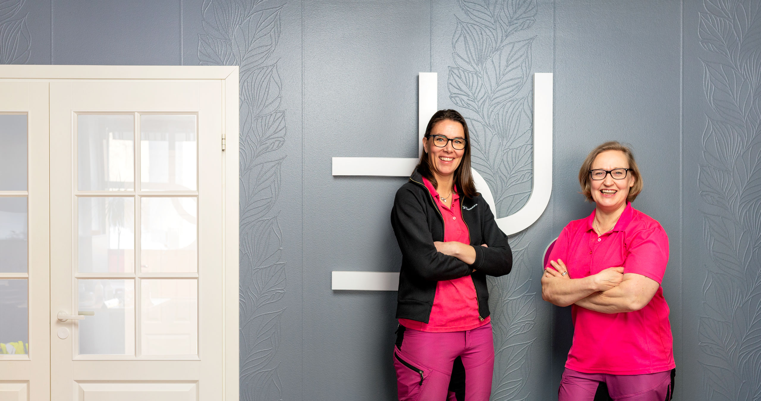 Duo siivouspalvelut Oy:n perustajat, Mari Männistö ja Sanna Kananen seisovat hymyillen kauniin harmaan seinän edessä, jossa on ruutuikkunallinen valkoinen ovi