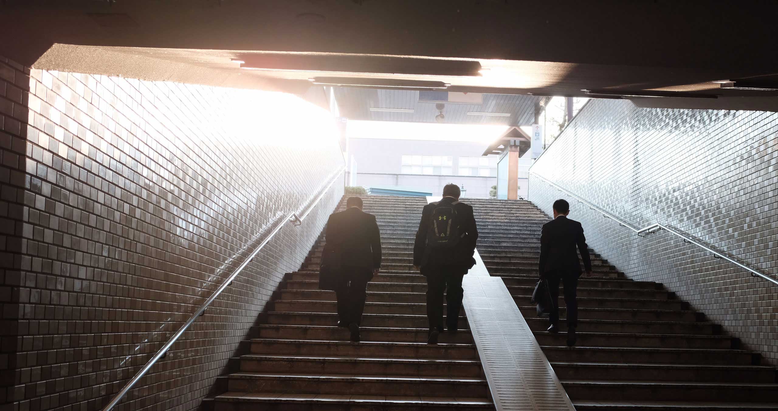 Kolme liikemiestä nousee metrokäytävän portaita pitkin kohti päivänvaloa.